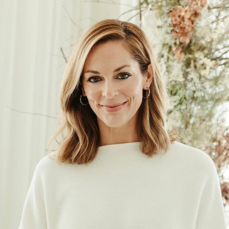 A woman with shoulder-length blonde hair wearing a cream-colored ribbed knit sweater, standing in front of a soft-focus floral background.